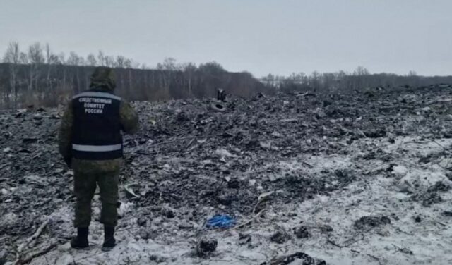 Detritos pretos e cinza espalhados na neve após a queda do avião Ilyushin-76.  Um investigador está parado de lado, olhando a cena. 