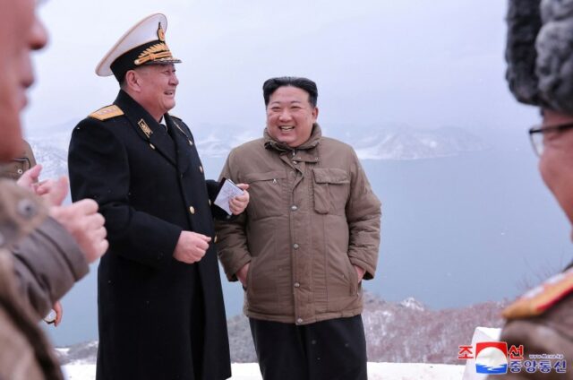 Kim Jong Un sorrindo enquanto conversa com oficiais militares.  Parece frio.  Ele está com as mãos nos bolsos.