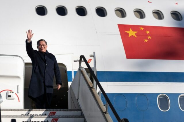 Li Qiang no topo da escada ao descer de um avião da Air China em Zurique.  Ele está vestindo um casaco preto e acenando.