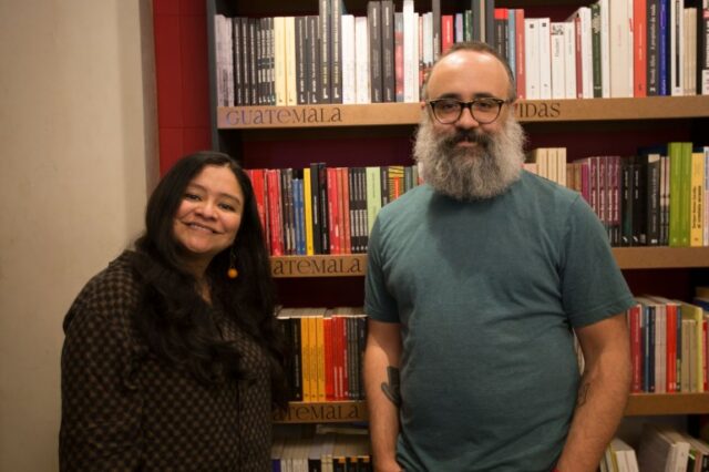 Duas pessoas ficam lado a lado em frente a uma estante de livros: um homem e uma mulher, o primeiro vestindo uma camiseta cinza e a segunda vestindo preta.