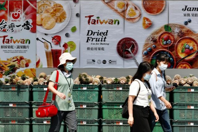 Pessoas passando por caixas de abacaxis.  Cartazes na parede atrás anunciam frutas tropicais de Taiwan