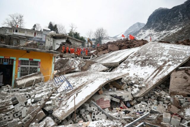 Equipes de resgate em uniformes laranja procuram sobreviventes nos escombros de edifícios desabados.  Um pequeno número de casas permanece de pé.  Algumas paredes caíram e estão cobertas de neve.