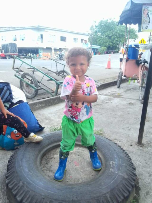 Emiliannys está no centro de um pneu caído no chão.  Ela usa botas de chuva azuis, calça verde e uma camisa rosa.