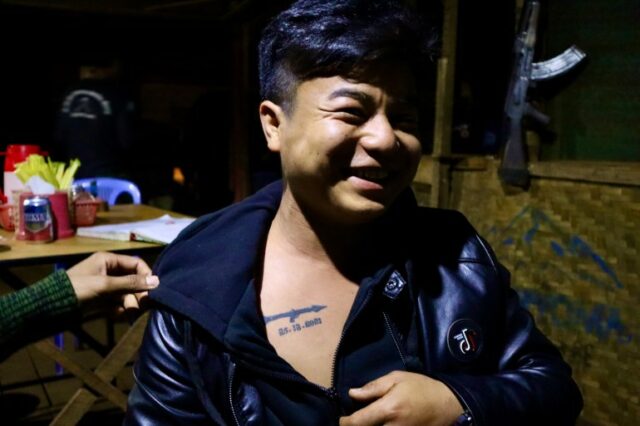     Lutador do Demoso PDF exibe tatuagem comemorativa da data em que foi ferido por um RPG militar.  Ele sorrindo e puxando a jaqueta para mostrar a tatuagem.