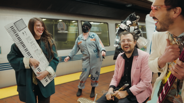 Membros da banda carregando um teclado de papel machê e um baixo elétrico sorriem uns para os outros ao lado de um baterista segurando um batedor na plataforma de uma estação de metrô, com uma dançarina parada perto de um trem.