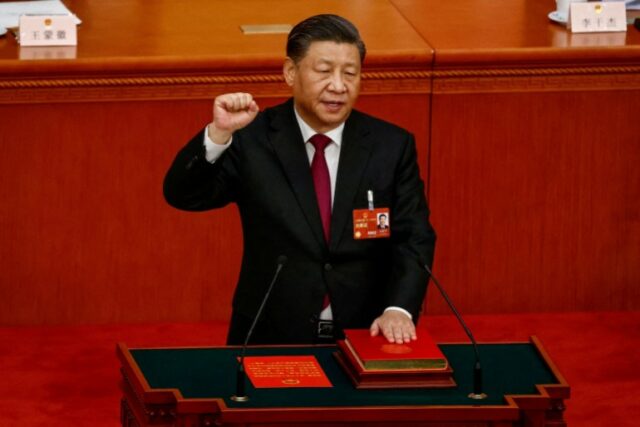 O presidente chinês, Xi Jinping, presta juramento de posse.  Ele tem a mão esquerda sobre a constituição chinesa e a direita em punho. Ele está no Grande Salão do Povo.