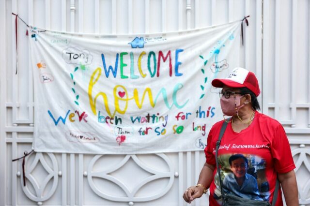 Um torcedor vestindo uma camiseta vermelha com a foto de Thaksin na frente e um boné vermelho e branco, em frente a uma faixa que diz: 'Bem-vindo ao lar.  Estávamos esperando por isso há tanto tempo’.