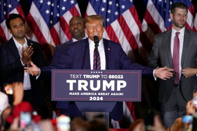 Donald Trump abre os braços atrás de um pódio estampado com seu nome.  Atrás dele estão apoiadores políticos e uma linha de bandeiras dos EUA.