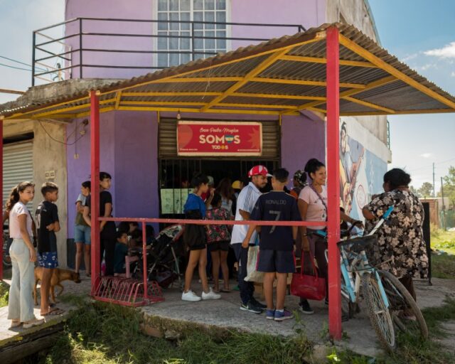 Uma fila de pessoas espera do lado de fora de um refeitório localizado em um prédio roxo de vários andares com toldo.