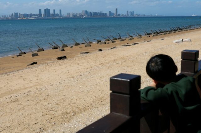 Uma criança olha para os arranha-céus de Xiamen da praia de Kinmen.  A praia é arenosa e o céu é azul.