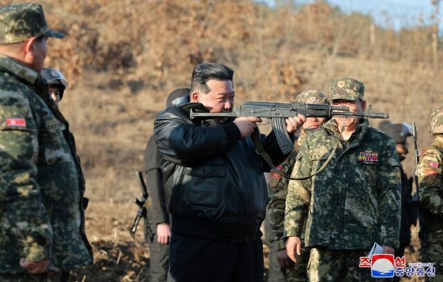Kim Jong Un segurando uma arma e olhando para o cano.  Ele está vestindo uma jaqueta de couro preta.  Seu cabelo está penteado para trás.  Há tropas em uniformes de combate atrás dele.