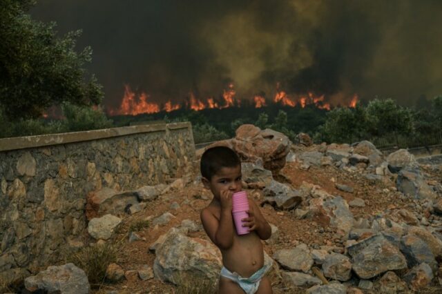 Uma criança está no quintal de sua casa enquanto um incêndio queima, na aldeia de Agios Charalampos, perto de Atenas, em 18 de julho de 2023. - A Europa se prepara para novas altas temperaturas em 18 de julho de 2023, sob uma onda de calor implacável e incêndios florestais que devastaram áreas do Hemisfério Norte, forçando a evacuação de 1.200 crianças perto de uma estância balnear grega.  As autoridades de saúde soaram alarmes desde a América do Norte até à Europa e Ásia, instando as pessoas a manterem-se hidratadas e a protegerem-se do sol escaldante, num duro lembrete dos efeitos do aquecimento global.  (Foto de Aris MESSINIS/AFP)