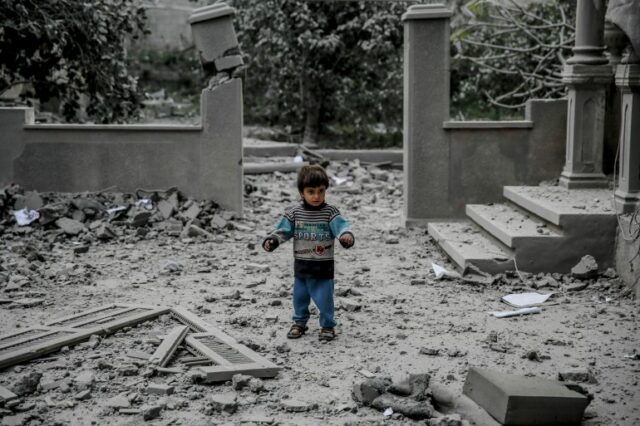 uma criança pequena está em frente a um prédio destruído coberto de poeira cinza