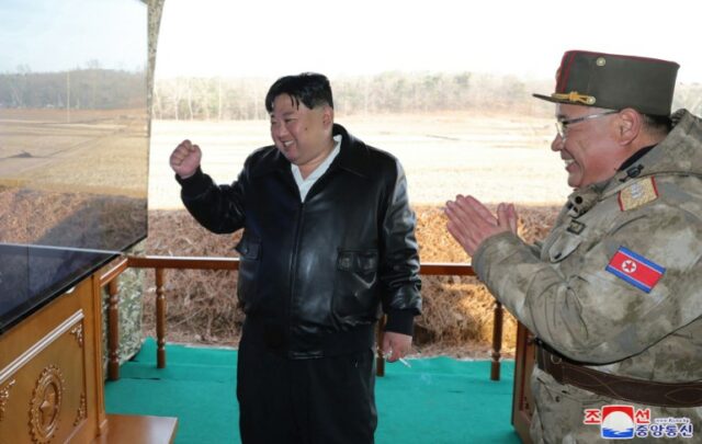 Kim Jong Un dá um soco no ar enquanto assiste a uma tela.  Um oficial militar com ele parece estar batendo palmas de entusiasmo.  Ambos estão sorrindo.