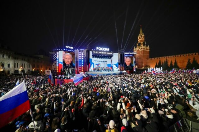 O presidente russo, Vladimir Putin, em evento na Praça Vermelha.  Seu rosto também aparece em telões ao lado do palco.  Parte da multidão agita bandeiras russas.  O Kremlin está em segundo plano.