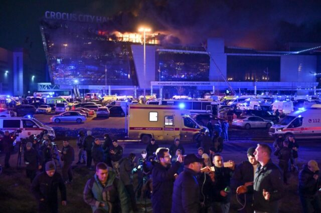 Pessoas do lado de fora do Crocus Concert Hall após o ataque.  As chamas sobem do topo do edifício.  Os veículos de emergência estão no estacionamento abaixo.