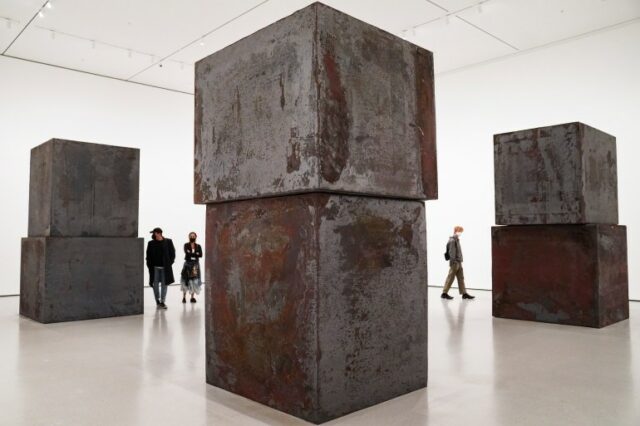 As pessoas que andam pelo Richard Serra trabalham 'Igualdade'.  Eles são ofuscados pelos imponentes blocos de aço enferrujados 
