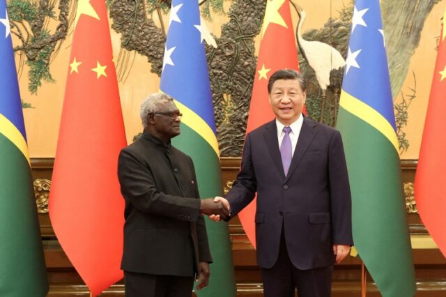 Manasseh Sogavare encontra-se com Xi Jinping em Pequim.  Eles estão apertando as mãos.  Xi está sorrindo.  As bandeiras dos seus dois países estão atrás deles.