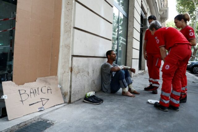 Trabalhadores da Cruz Vermelha verificam um sem-teto durante uma onda de calor na Itália.  O homem está sentado na rua, encostado a uma parede.  Ele tirou os sapatos e está descalço.  Os três funcionários da Cruz Vermelha estão uniformizados e conversam com ele. 