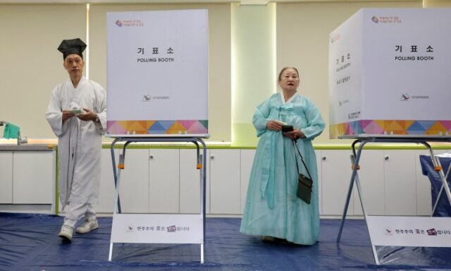 Um professor da aldeia e a sua família nas assembleias de voto.  Eles usam trajes tradicionais e ficam ao lado das cabines de votação.