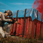 Imagem de PUBG, mostrando um jogador se escondendo atrás de uma caixa com um rifle de assalto.  Cena rural com uma casa antiga atrás.