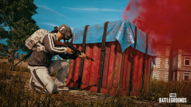 Imagem de PUBG, mostrando um jogador se escondendo atrás de uma caixa com um rifle de assalto.  Cena rural com uma casa antiga atrás.