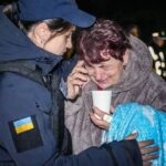 Um policial conforta uma mulher idosa em Odesa depois que um ataque russo destruiu mais de uma dúzia de apartamentos.  A mulher mais velha está ao telefone segurando uma xícara de chá.  Ela está soluçando.  A policial, uma mulher mais jovem, dá-lhe um abraço e parece preocupada.