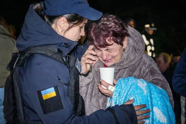 Um policial conforta uma mulher idosa em Odesa depois que um ataque russo destruiu mais de uma dúzia de apartamentos.  A mulher mais velha está ao telefone segurando uma xícara de chá.  Ela está soluçando.  A policial, uma mulher mais jovem, dá-lhe um abraço e parece preocupada.