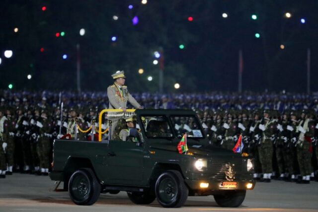 Min Aung Hlaing inspecionando tropas no desfile anual do Dia das Forças Armadas.  Ele está na traseira de um veículo semelhante a um jipe ​​​​de capota aberta.  É noite.