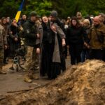 Dois homens apoiam uma mulher idosa no funeral do paramédico do exército ucraniano Nazarii Lavrovskyi.  As pessoas estão reunidas atrás.  alguns de uniforme e outros de preto.  Um deles tem a bandeira da Ucrânia.  Há uma pilha de terra à direita da imagem.