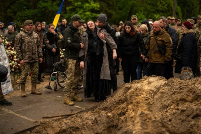 Dois homens apoiam uma mulher idosa no funeral do paramédico do exército ucraniano Nazarii Lavrovskyi.  As pessoas estão reunidas atrás.  alguns de uniforme e outros de preto.  Um deles tem a bandeira da Ucrânia.  Há uma pilha de terra à direita da imagem. 