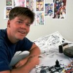 Um jovem de pele clara se apoia em uma mesa de artista, com desenhos em preto e branco e uma edição da revista em quadrinhos