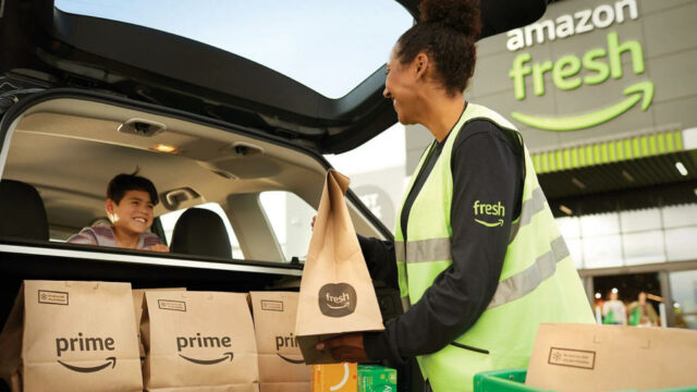 Um funcionário da Amazon Fresh com um colete verde neon carrega mantimentos Prime Fresh em um hatchback com uma criança sorrindo no banco de trás.  A pessoa que carrega as compras sorri de volta. 