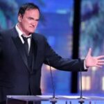 Quentin Tarantino, um homem de pele clara e cabelo preto curto, usa smoking e fala em frente a um estrado com dois microfones saindo dele.  Ele estende os braços, gesticulando, diante de um fundo de um falso horizonte com palmeiras.