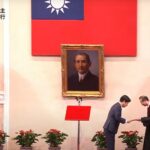 William Lai Ching-te durante sua cerimônia de inauguração.  Há uma bandeira de Taiwan na parede e um retrato de Sun Yat-sen, fundador da República da China.  com grandes exibições de orquídeas cor de rosa em ambos os lados.  Lai está fazendo uma reverência ao receber um documento de um funcionário.