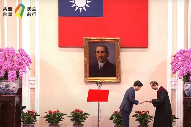 William Lai Ching-te durante sua cerimônia de inauguração.  Há uma bandeira de Taiwan na parede e um retrato de Sun Yat-sen, fundador da República da China.  com grandes exibições de orquídeas cor de rosa em ambos os lados.  Lai está fazendo uma reverência ao receber um documento de um funcionário.