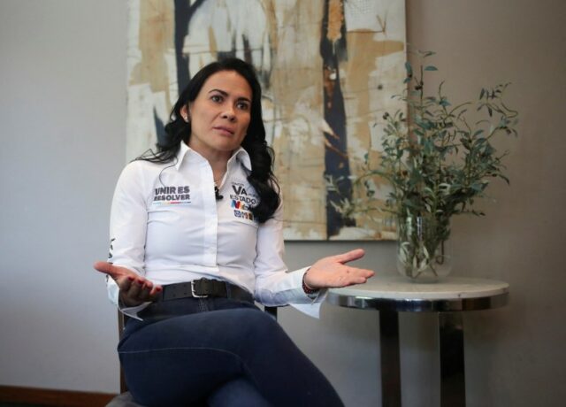 Alejandra del Moral está sentada para uma entrevista, com as mãos abertas em gestos enquanto fala.  Uma pequena mesa e uma pintura são visíveis atrás dela.