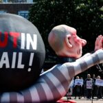 Uma efígie gigante de Vladimir Putin em traje listrado de prisão.  Suas mãos estão acorrentadas e pintadas de vermelho.  'Putin Jail' está escrito em uma bola preta nas costas do boneco.  Os manifestantes estão reunidos nas proximidades.  Eles seguram cartazes pedindo a prisão de Putin.