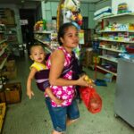 Uma mulher da etnia Chin de Mianmar dentro de uma loja de artigos diversos na Malásia.  Ela tem seu filho amarrado às costas em um sarongue.  Ela está carregando um pequeno saco plástico.