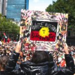 Uma mulher segurando um cartaz em um comício.  Diz que a Austrália Branca tem uma História Negra em uma bandeira indígena, e há marcas de mãos pretas nela.  Há um mar de pessoas à sua frente.