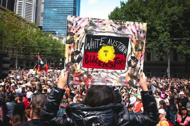 Uma mulher segurando um cartaz em um comício.  Diz que a Austrália Branca tem uma História Negra em uma bandeira indígena, e há marcas de mãos pretas nela.  Há um mar de pessoas à sua frente.