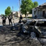 A polícia ucraniana inspeciona carros destruídos por uma bomba russa em Zolochiv.  Os carros estão estacionados ao lado da estrada, mas queimados.  O telhado de um prédio próximo a eles foi destruído e as janelas quebradas