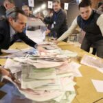 Os funcionários eleitorais esvaziam uma urna sobre uma mesa.  Há uma grande pilha de boletins de voto que eles estão organizando.