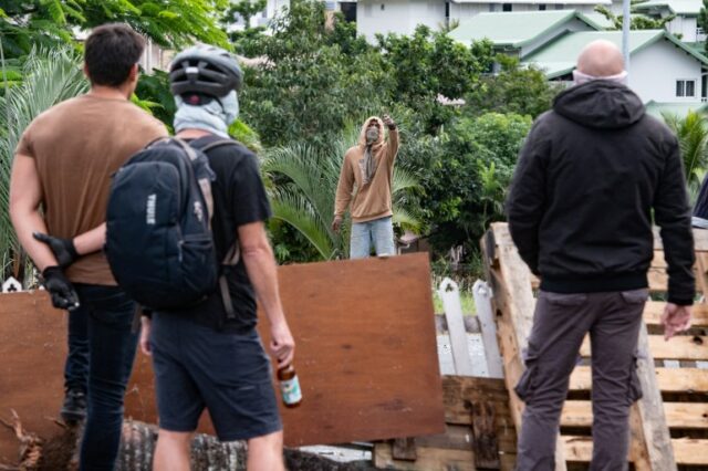 Pessoas frente a frente numa barricada em Nouméa.  Há três homens de costas para a câmera e outro homem parado do outro lado.  O homem está mascarado e usando um moletom.  Ele está gesticulando em direção a eles.