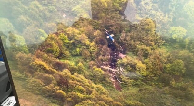 Uma área negra queimada na encosta de uma montanha arborizada.  A cauda azul e branca de um helicóptero pode ser vista.