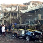 Cena dos atentados de Bali em 2002. Policiais inspecionam as ruínas dos edifícios devastados.  Outros estão assistindo.  Um carro destruído está na frente.  Tem fita policial amarela ao redor.