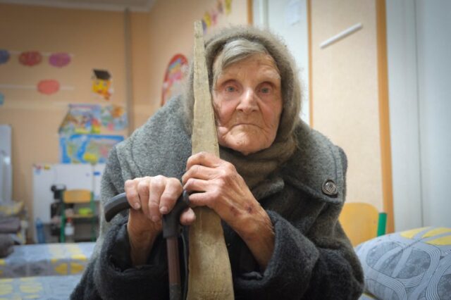 Lídia Stepanovna.  Ela está sentada em uma cama com um xale na cabeça e apoiada em uma bengala.