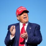Donald Trump bate palmas contra um céu azul claro, usando um boné MAGA vermelho.
