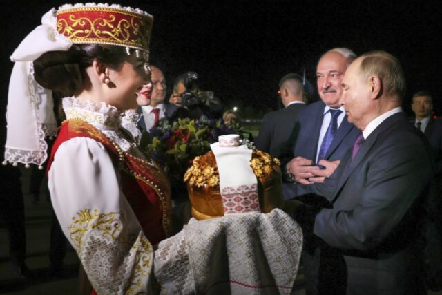 Alexander Lukashenko dá as boas-vindas a Vladimir Putin na Bielorrússia.  Há uma mulher em traje tradicional à esquerda segurando um presente.  Uma mulher atrás dela tem um buquê de flores.  Lukashenko e Putin estão à direita.