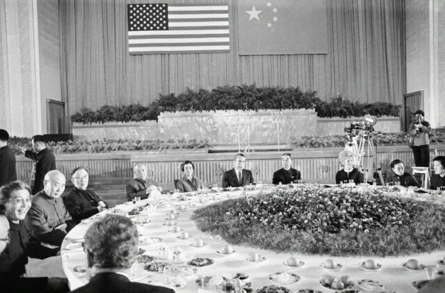 O presidente dos EUA, Richard, e a primeira-dama Pat Nixon em uma enorme mesa de banquete em sua visita à China em 1972. O primeiro-ministro chinês, Zhou Enlai, está do outro lado da mesa.  As bandeiras da China e dos EUA estão expostas na parede atrás delas